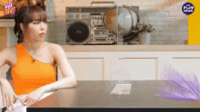 a woman in an orange top sits at a table in front of a boombox that says the play on it