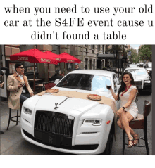 a man and a woman sit in front of a white car with a table on the hood