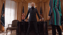 a man in a suit stands in front of a desk in a room with american flags in the background