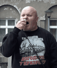 a bald man wearing a black shirt that says " dogodine y pri3pby " is shouting into a microphone