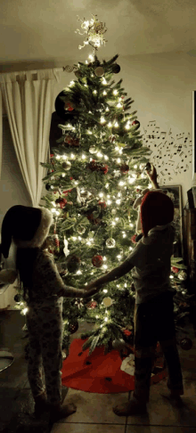 two children wearing santa hats decorate a christmas tree in a living room