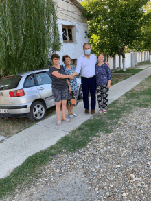 a man in a mask shakes hands with two women in front of a silver car with the license plate r-b-jx