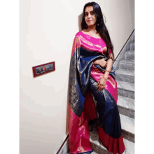 a woman in a pink and blue saree is standing in front of stairs