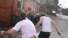 two men are running down a street in front of a brick building . one of the men is wearing a white tank top .