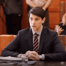 a man in a suit and tie is sitting at a table with his hands folded and a glass of water .