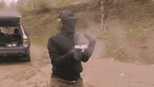 a man wearing a mask and goggles is holding a gun in front of a car on a dirt road .