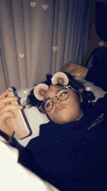 a person wearing glasses and bear ears laying on a bed looking at their phone