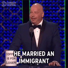 a man in a tuxedo stands at a podium with a mug that says " he married an immigrant "