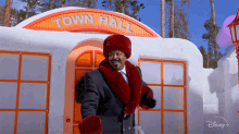 a man in a suit and red fur coat is standing in front of a snowy building that says town hall