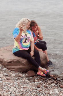 two women sit on a rock near the water and one has a tie dye shirt that says niagara falls