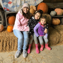 a woman and two little girls are sitting on a bale of hay with pumpkins in the background