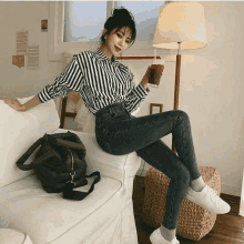 a woman in a striped shirt and jeans is sitting on a couch holding a cup of coffee .