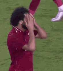 a man in a maroon shirt is covering his ears with his hands while standing on a soccer field .