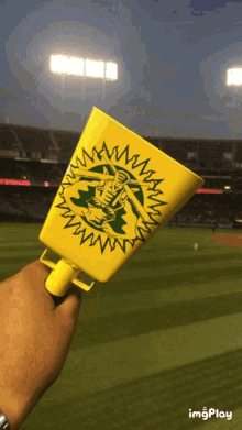 a person is holding a yellow cup that says ' oakland ' on it in front of a baseball field