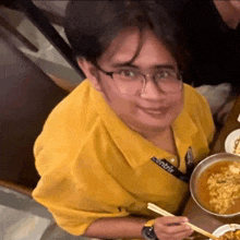 a man in a yellow shirt and glasses is sitting at a table with chopsticks and a bowl of food .