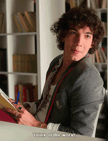 a young man with curly hair is sitting at a desk with a book in his hand and a caption that says think of the worst