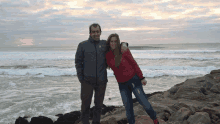 a man and a woman are posing for a picture on the beach and the man is wearing a grey jacket with the letter t on it