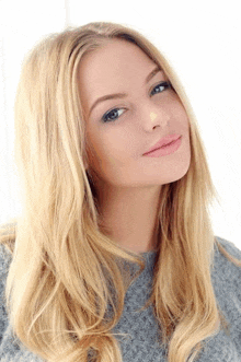 a woman with long blonde hair is wearing a grey sweater and smiling at the camera .