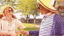 two women are standing next to each other in a park holding hands . one of the women is wearing a straw hat .