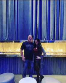 a man and a woman pose in front of a blue curtain with the man wearing a freestyle shirt