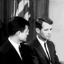 two men in suits and ties are sitting next to each other in front of an american flag in a black and white photo .