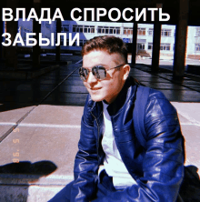 a young man wearing sunglasses and a blue jacket sits on a sidewalk in front of a building