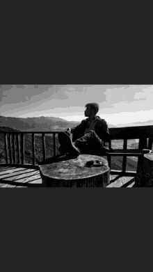 a black and white photo of a man sitting on a wooden bench overlooking a mountain .