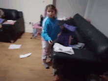 a little girl in a blue jacket stands in front of a black couch