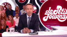 a man in a suit and tie sits at a table in front of a sign that says le petit journal