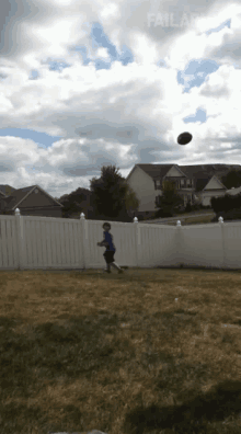 a boy in a blue shirt is running towards a white fence while a football is flying in the air