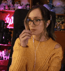 a woman wearing glasses and a yellow sweater is sitting in front of a shelf with stuffed animals on it