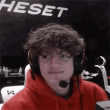 a young man wearing a red hoodie and headphones is sitting in front of a sign that says heset .