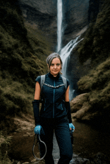 a woman stands in front of a waterfall wearing gloves