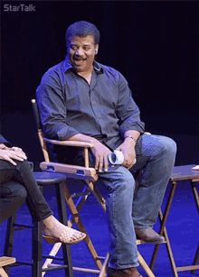 a man in a black shirt sits in a director 's chair on a stage with startalk written on the bottom