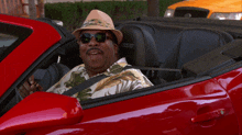 a man wearing a hat and sunglasses is driving a red car with the word ford on the seat