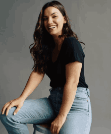 a woman in a black shirt and blue jeans is sitting on a stool