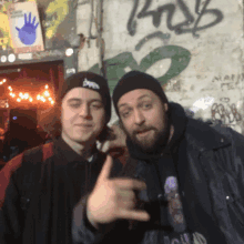 two men are posing for a picture in front of a wall with graffiti on it that says ' bob '