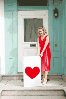 a woman in a red dress holds a white box with a red heart on it in front of a blue door