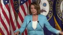 a woman in a blue jacket stands in front of a microphone in front of a house of representatives flag