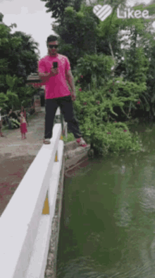 a man in a pink shirt is standing on a bridge over a body of water and looking at his phone