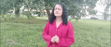 a woman in a pink shirt is standing in a field with her hands folded .