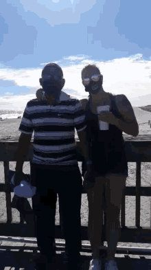 a man and a woman wearing face masks are standing next to each other on a pier at the beach .