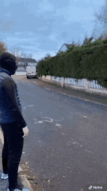 a man wearing a black mask is standing on the side of a road next to a white van and a hedge