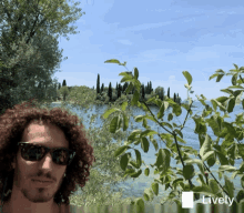 a man with curly hair wearing sunglasses stands in front of a body of water