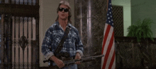 a man in a plaid shirt is holding a shotgun in front of an american flag .