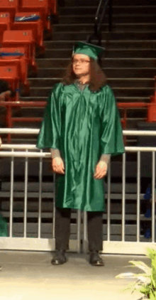 a man in a green graduation cap and gown stands in front of a fence