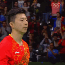 a man wearing a red shirt with a dragon on it stands in front of a crowd at the olympics