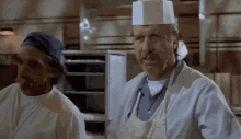 a man in a chef 's hat is standing next to another man in an apron in a kitchen .