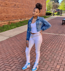 a young girl wearing a denim jacket , white jeans , and blue sneakers is standing on a brick sidewalk .