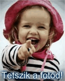 a little girl wearing a pink hat and a striped shirt is pointing at the camera .